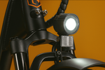 A black illuminated headlight with silver brackets on a matte black ebike. 