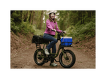 A woman on a trail in a forest sitting on a green cargo ebike with a small front basket and large rear basket wearing a pink jacket and white helmet.
